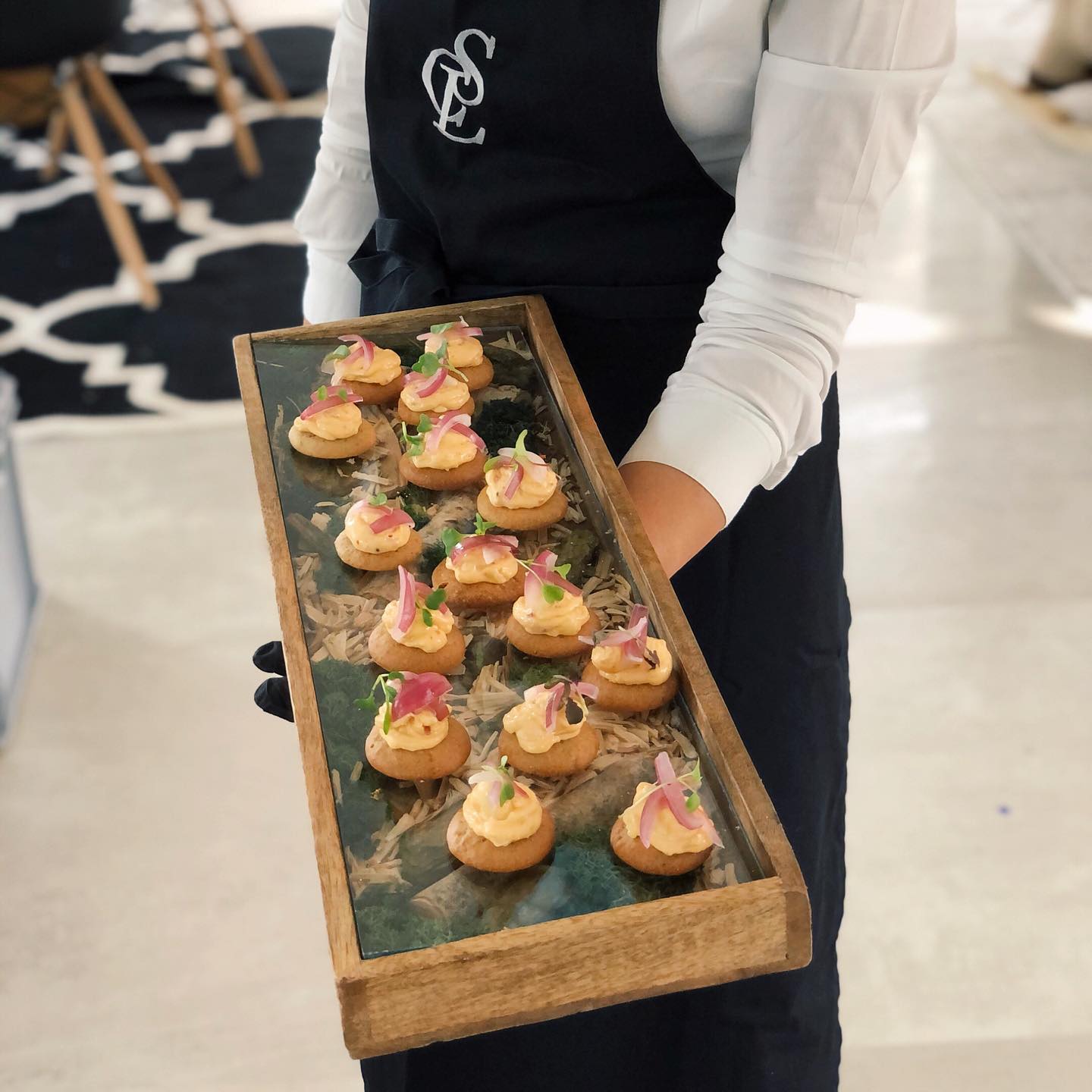 CSL Catering in München für Business und private Feiern 48 Ein CSL Catering Kellner serviert ein Flying buffet auf einem eigens kreierten Tablett mit Wald-Thema, sichtbar durch eine Glasscheibe auf einem hölzernen Rahmen.